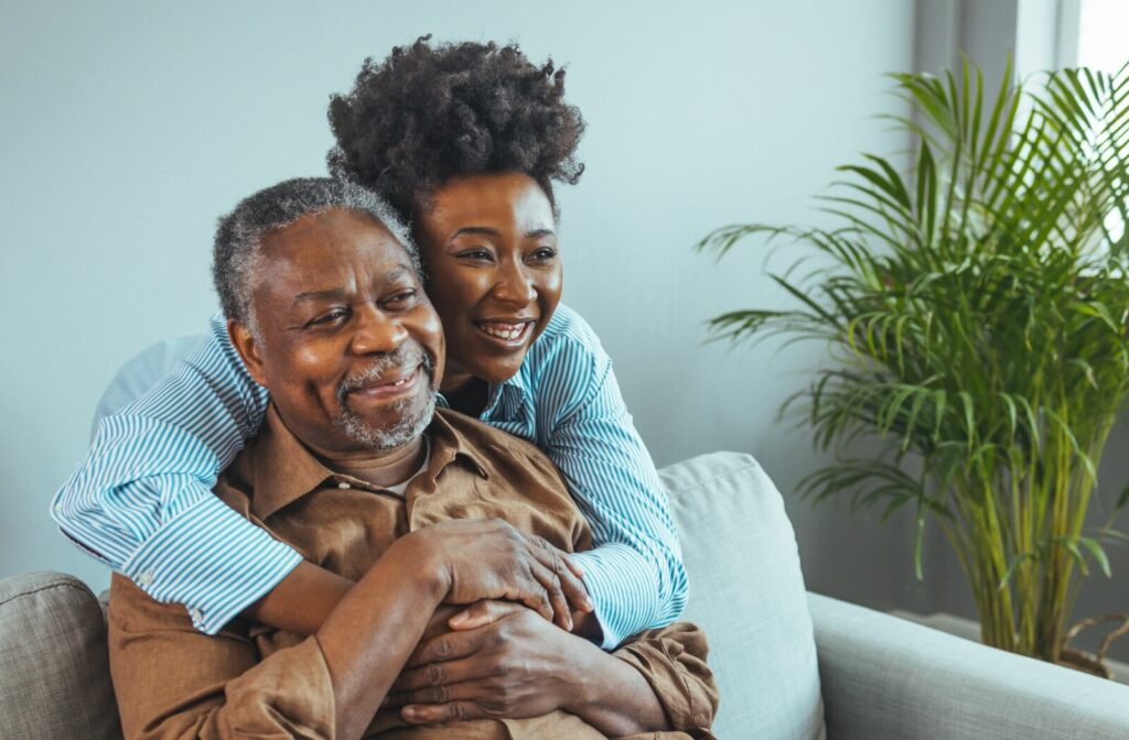 An adult daughter visits her father in his assisted living home, giving him a hug from behind where he sits on the couch.
