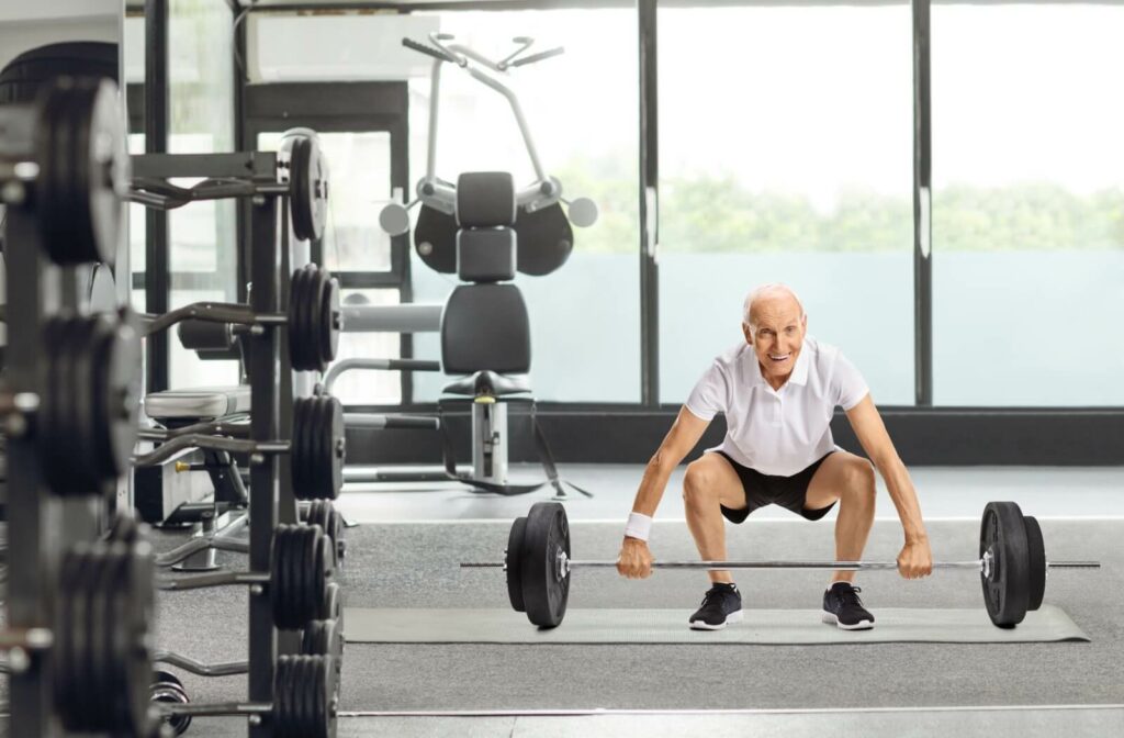 An older adult man bends his hips and knees to lift a barbell in a gym.