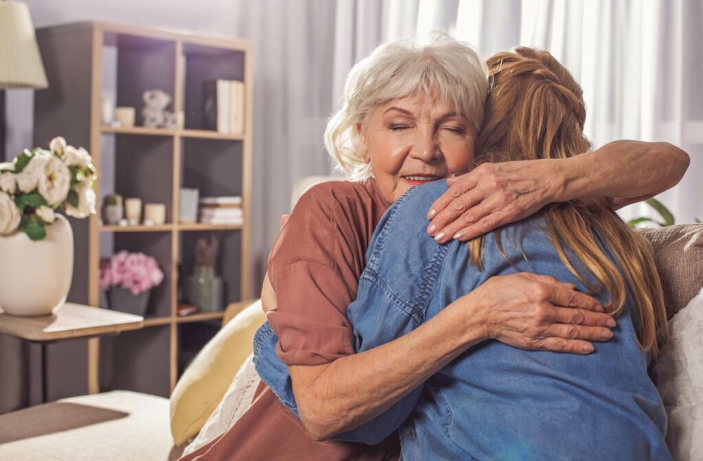 An adult daughter hugs her mother on the couch in her personal care home apartment.
