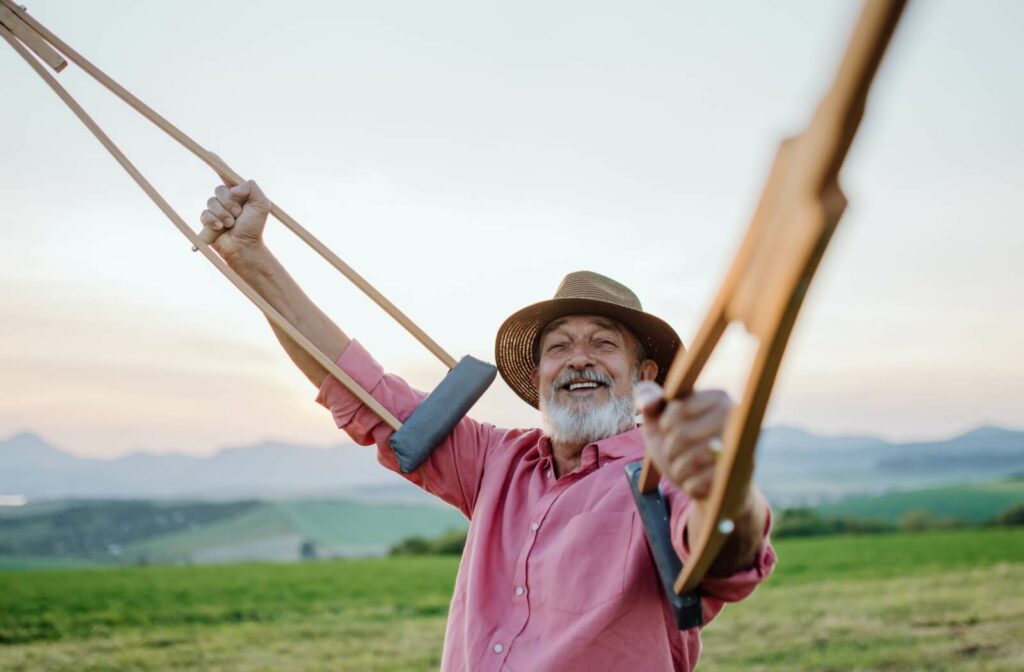A smiling older adult man lifts crutches in the air while on an outdoor walk.
