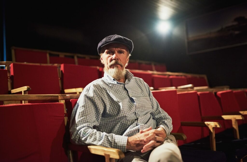 An older adult sits confidently in a cinema chair with their hands clasped in their lap.